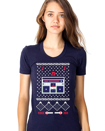 A navy blue women’s t-shirt displays a pixelated graphic reminiscent of a traditional Christmas sweater. At the center is a stylized boombox in white, red, and dark blue, surrounded by small dots, zigzag borders, and red musical notes. The design blends festive sweater aesthetics with 1980s hip-hop boombox culture.
