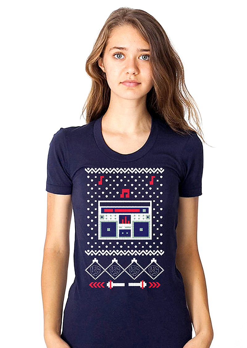 A navy blue women’s t-shirt displays a pixelated graphic reminiscent of a traditional Christmas sweater. At the center is a stylized boombox in white, red, and dark blue, surrounded by small dots, zigzag borders, and red musical notes. The design blends festive sweater aesthetics with 1980s hip-hop boombox culture.