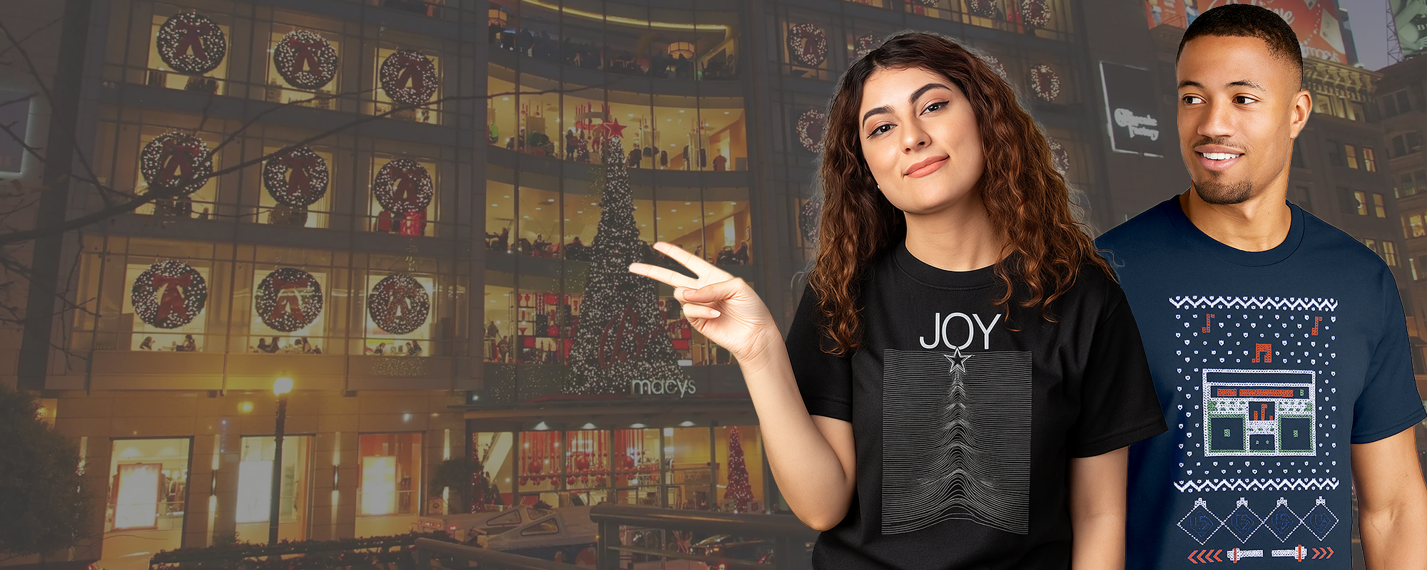 Two people wearing holiday graphic t-shirts — one reading ‘JOY’ with a minimalist line design inspired by the 80s music group Joy Division, and the other featuring an cross-stitch holiday sweater pattern — stand in front of a warmly lit department store decorated with Christmas wreaths and lights.
